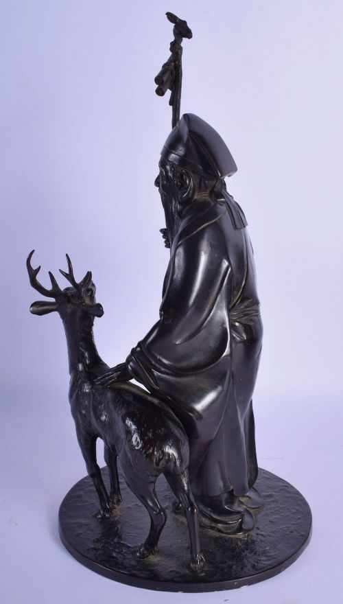 fig 1. A dark patinated bronze okimono sculpture depicting the Japanese deity Jurojin holding a tall staff tied with a scroll, standing beside a small stag on a circular bronze base.