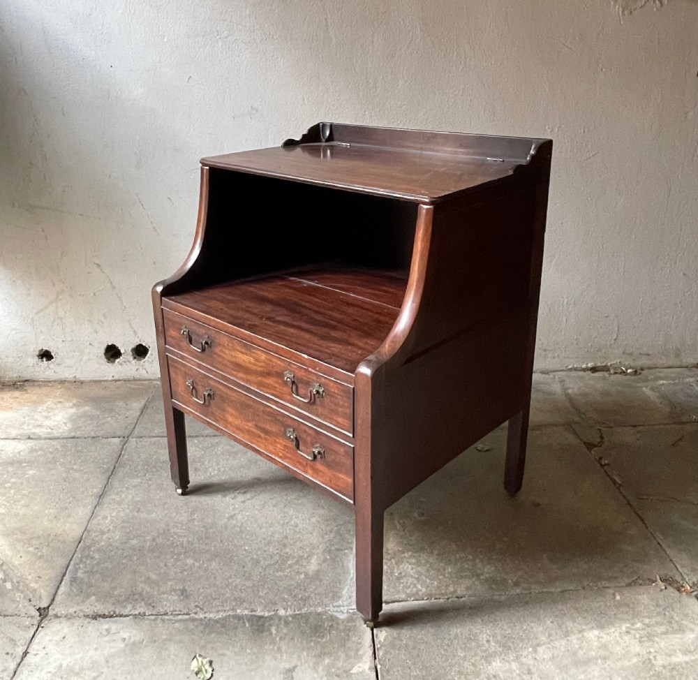 A George III dark mahogany bedside commode featuring an open upper shelf, a shaped tray top, two lower drawers with brass handles, and square legs on small brass castors.