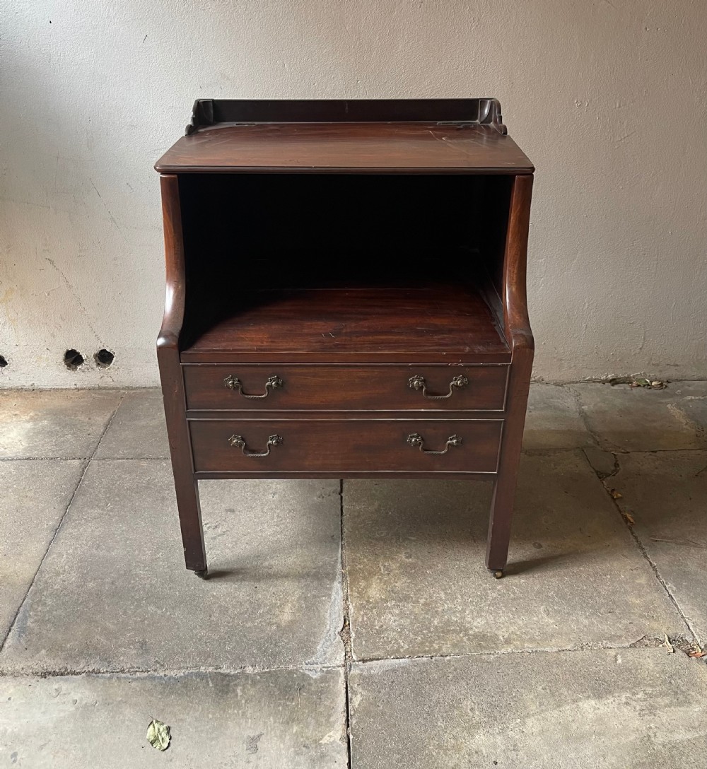 A George III dark mahogany bedside commode featuring an open upper shelf, a shaped tray top, two lower drawers with brass handles, and square legs on small brass castors. View II