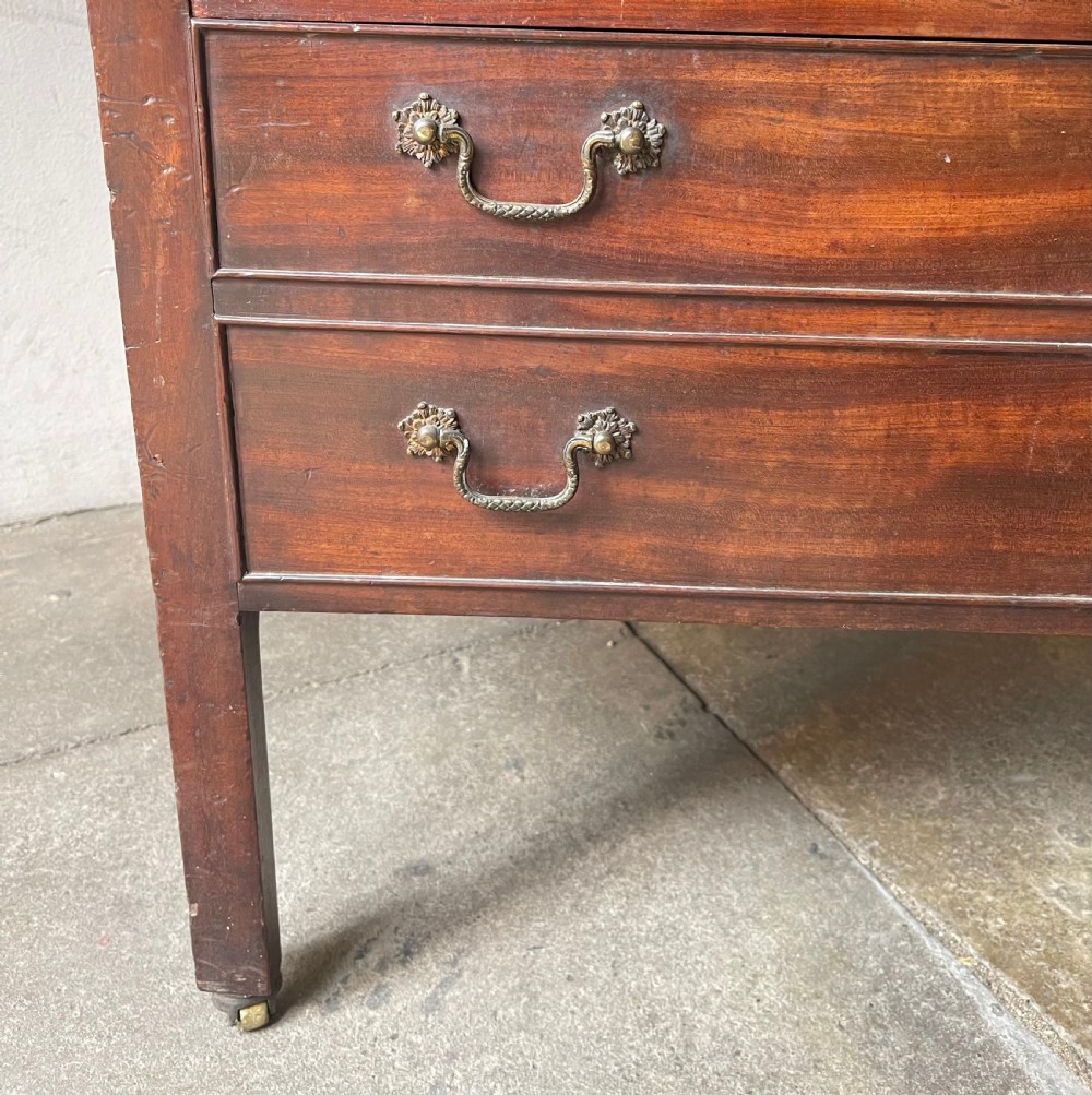 A George III dark mahogany bedside commode featuring an open upper shelf, a shaped tray top, two lower drawers with brass handles, and square legs on small brass castors. View V