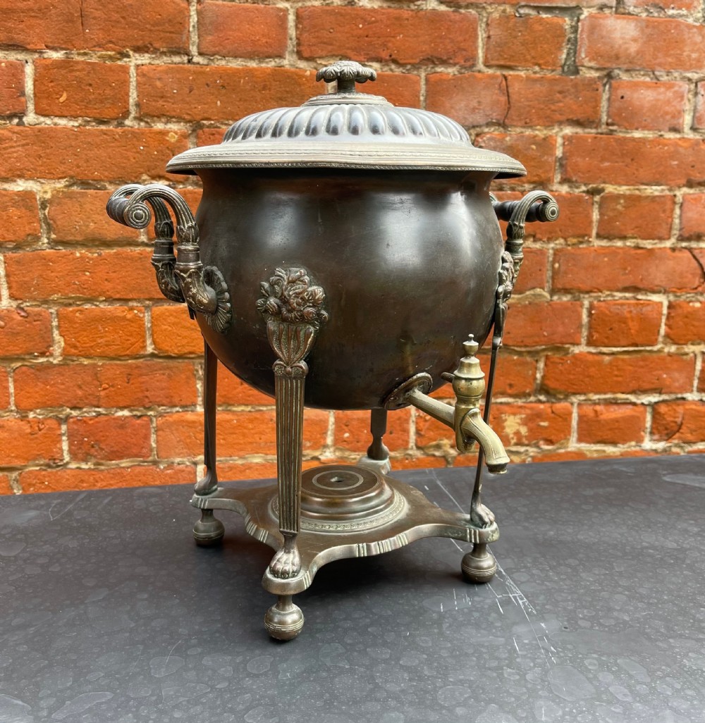 An antique Regency patinated metal tea urn featuring a fluted lid, twin scroll handles, a front brass spigot, and reeded legs resting on a shaped base. View II