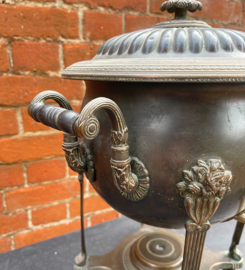 An antique Regency patinated metal tea urn featuring a fluted lid, twin scroll handles, a front brass spigot, and reeded legs resting on a shaped base. View IV