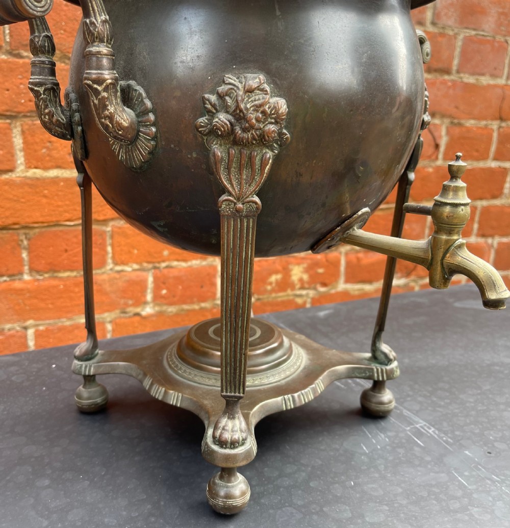 An antique Regency patinated metal tea urn featuring a fluted lid, twin scroll handles, a front brass spigot, and reeded legs resting on a shaped base. View V