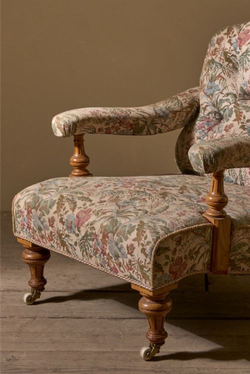fig 1. A 19th-century Victorian open armchair with floral pattern fabric upholstery, a deep-buttoned backrest, turned wooden front legs on brass castors, and padded armrests.