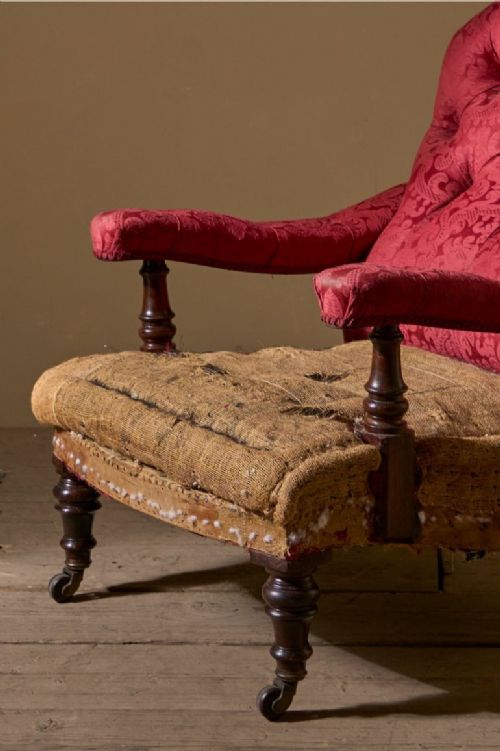fig 1. A 19th-century Victorian armchair featuring a tufted red damask buttoned backrest, open padded armrests on turned mahogany supports, an exposed hessian seat base, and turned wooden front legs with brass castors.