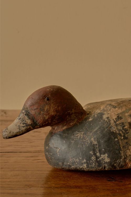 fig 2. An early 20th-century hand-carved wooden duck decoy with distressed red paint on the head and a weathered dark body resting on a wooden surface.