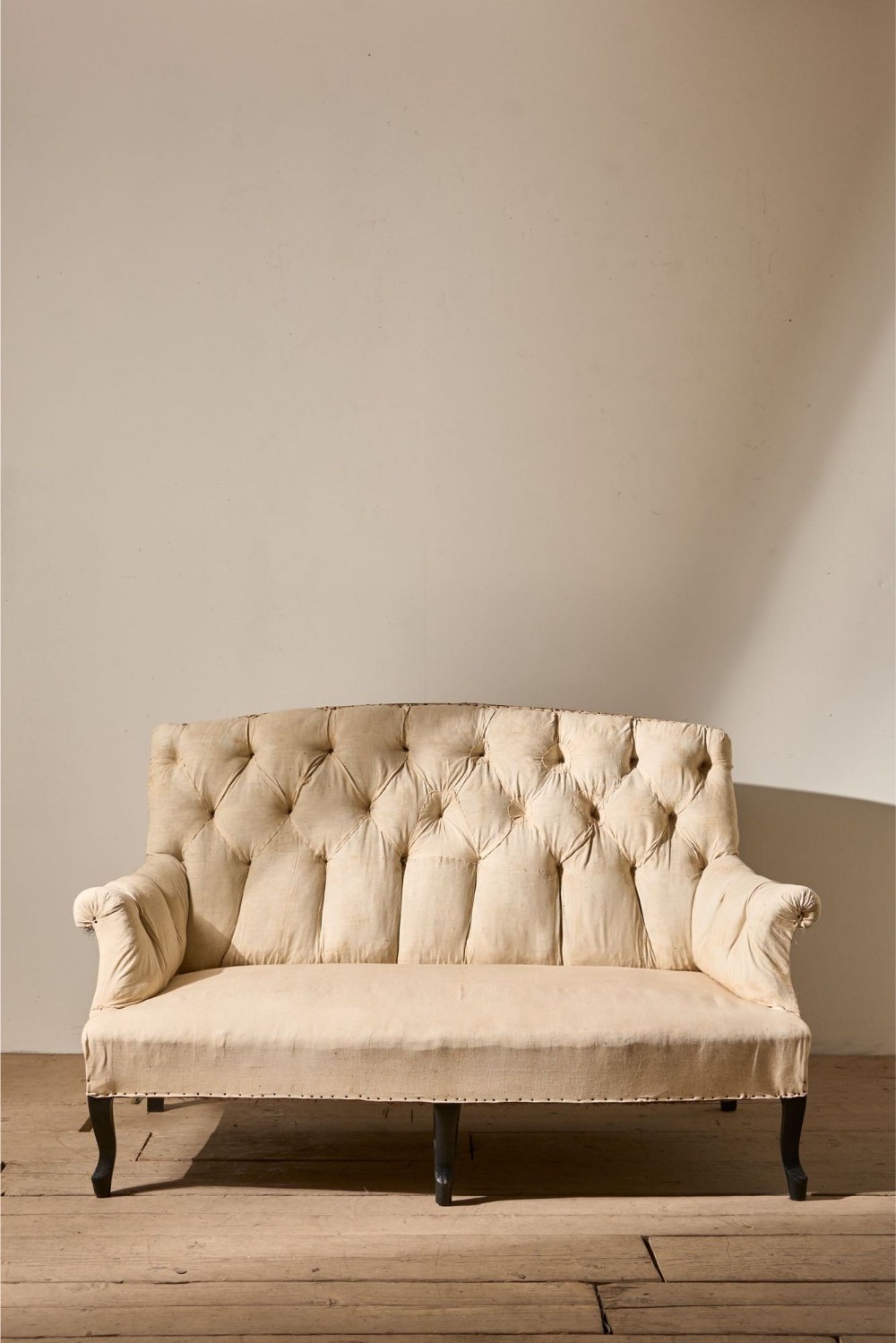 A small Napoleon III sofa featuring deep button-tufted cream upholstery, a high curved backrest, rolled arms, and black turned wooden legs.