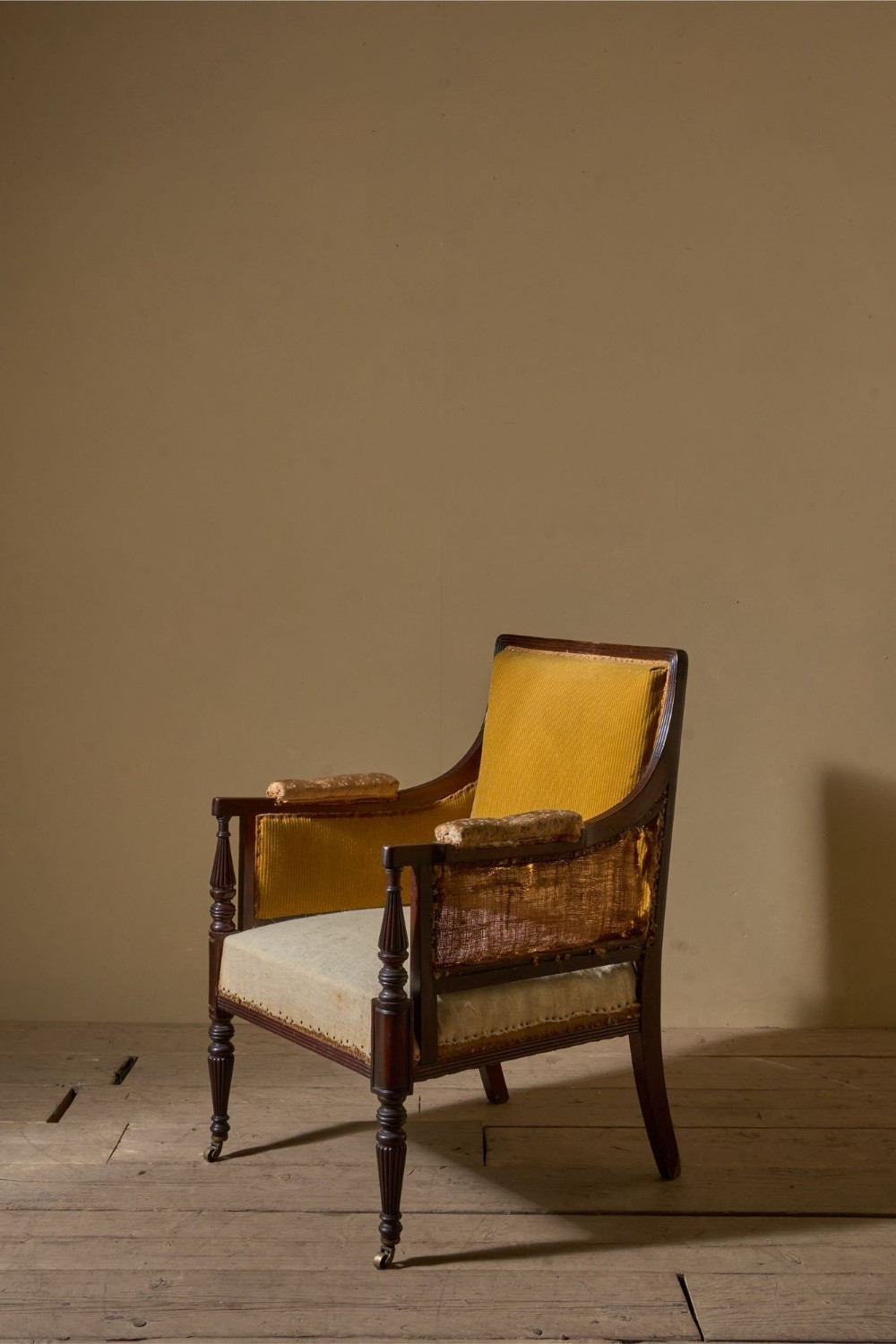 A 19th century mahogany library chair featuring reeded arms and legs, brass castors, exposed hessian sides, and yellow corduroy cushion fabric.
