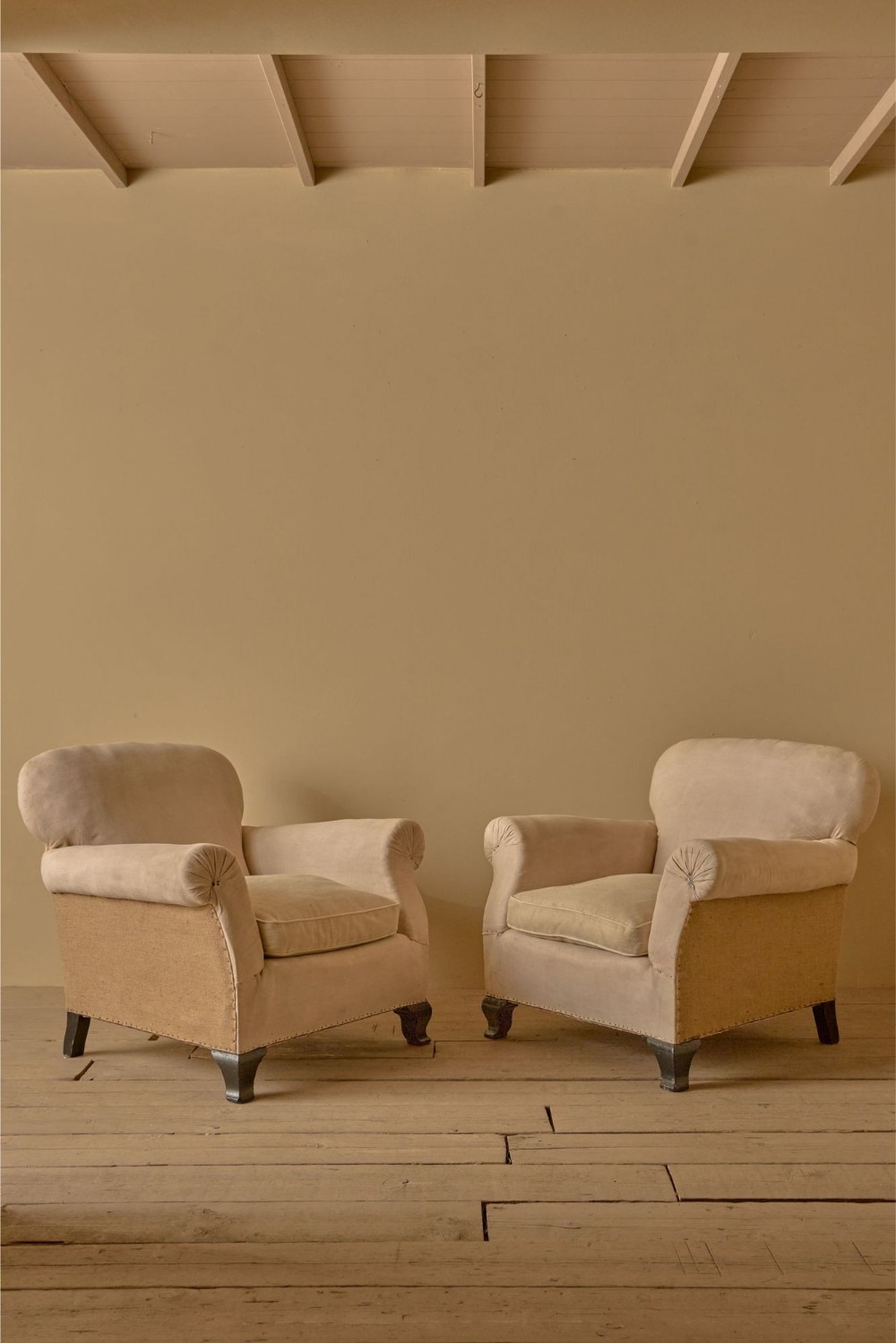 A pair of 1920s French club armchairs upholstered in neutral beige calico with exposed hessian lower panels and dark turned wooden feet.