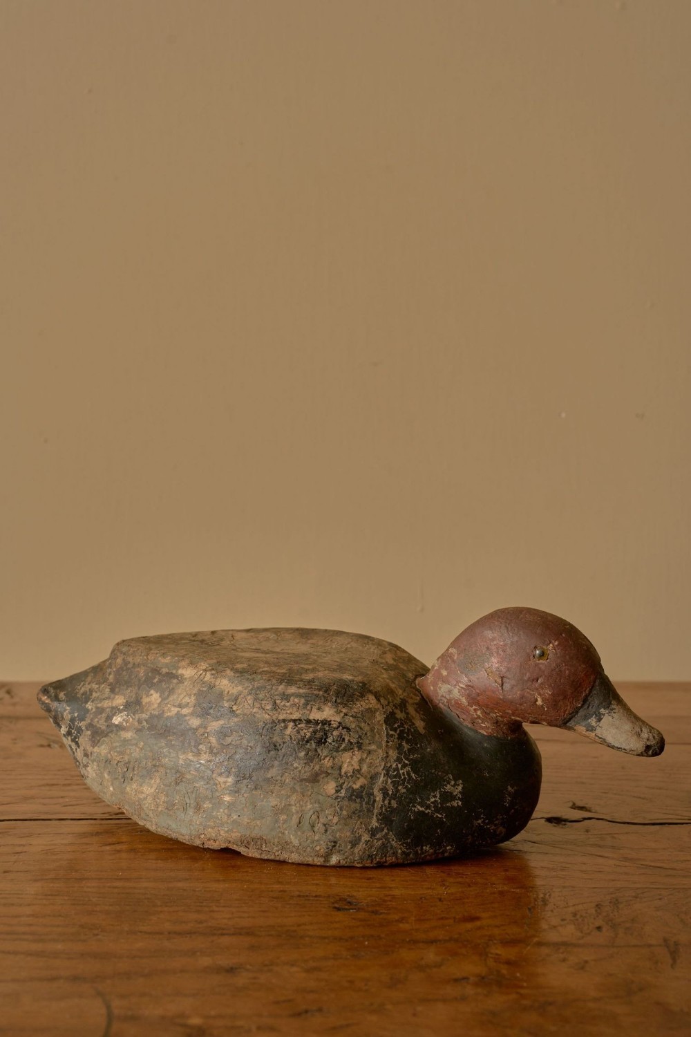 An early 20th-century hand-carved wooden duck decoy with distressed red paint on the head and a weathered dark body resting on a wooden surface.