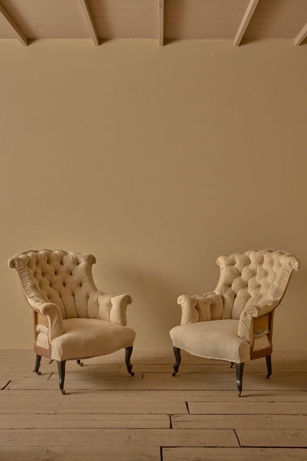 A pair of French 19th-century Napoleon III high-back armchairs upholstered in cream fabric with deep buttoning and dark legs on castors.