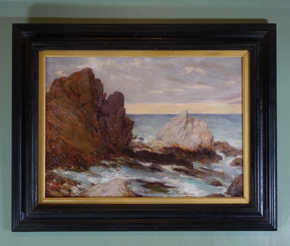 An early 20th-century impressionist oil painting depicting waves crashing against rocky coastal cliffs under cloudy grey skies, enclosed in a dark molded wooden frame with a gilded inner border.