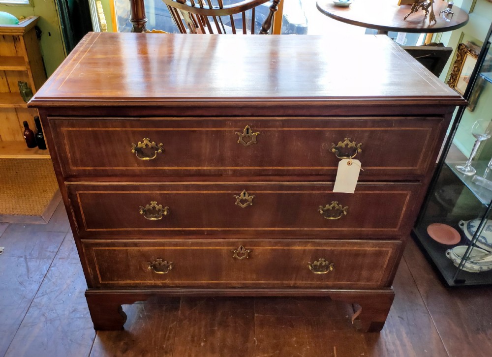 A late eighteenth-century mahogany chest of three drawers featuring fine boxwood stringing inlay, brass swan-neck handles, keyhole escutcheons, and bracket feet.