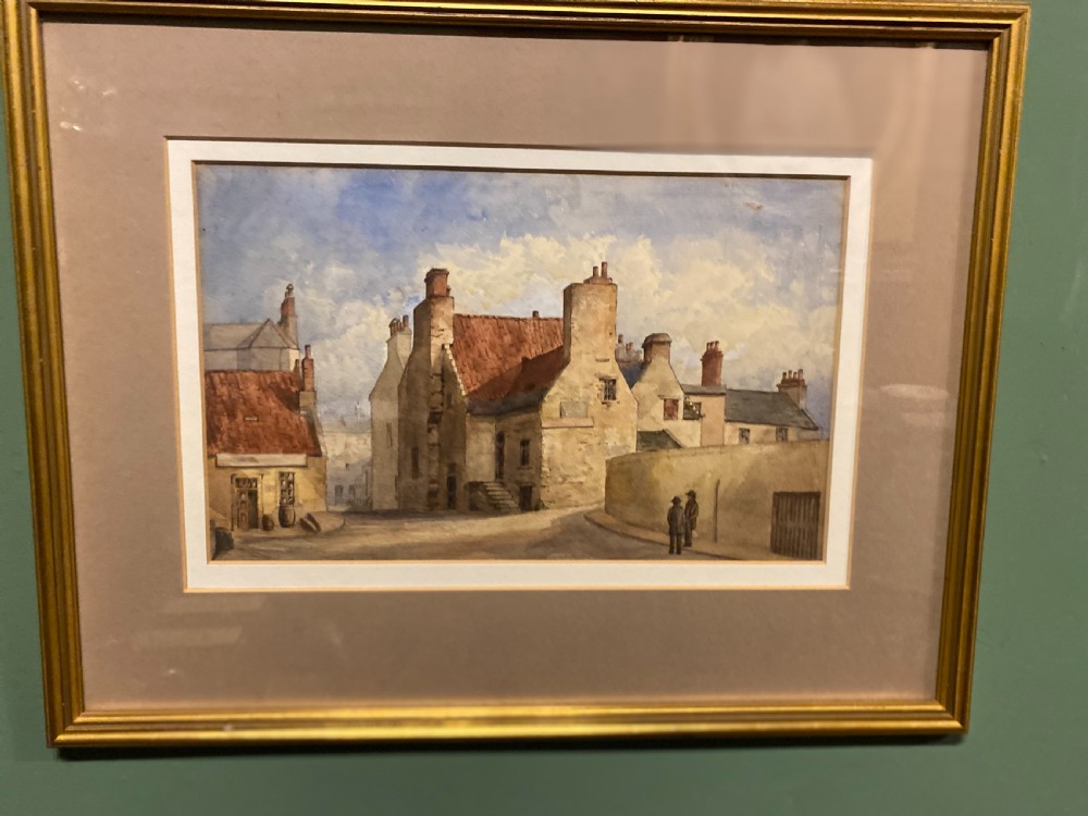 A framed watercolour painting depicting stone village houses with red-tiled roofs along a quiet street under a cloud-filled sky.