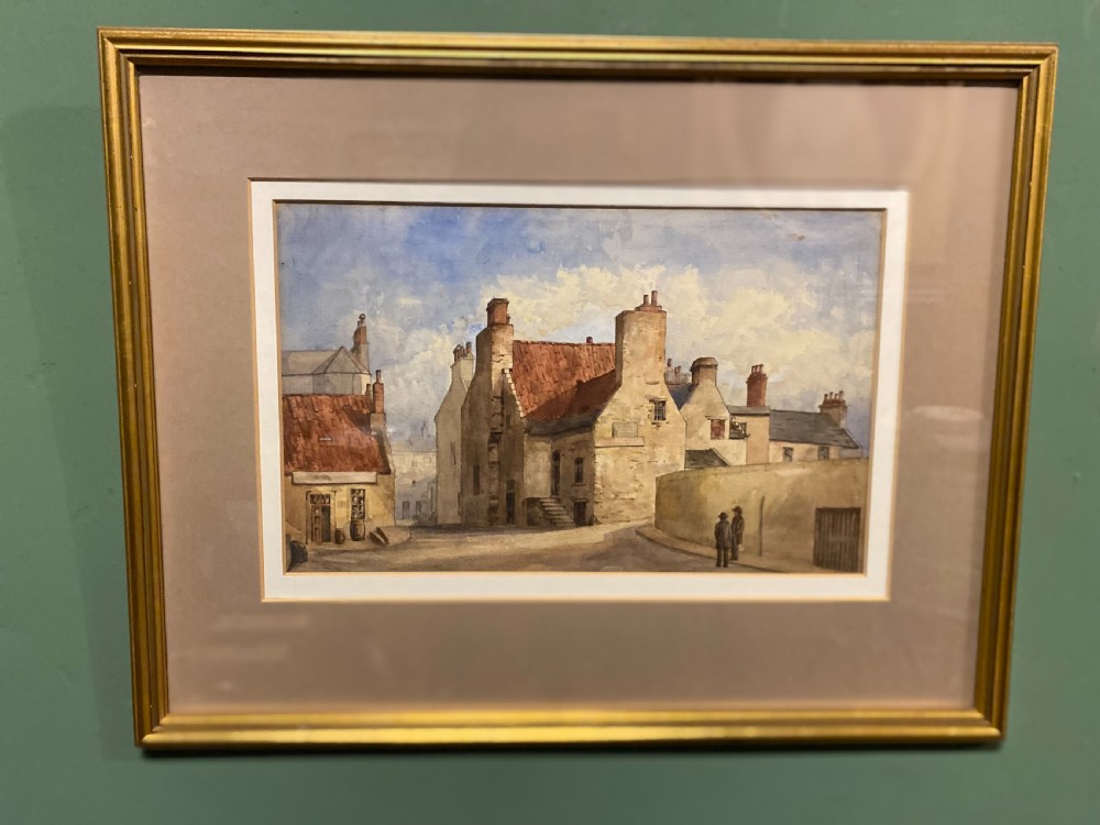 A framed watercolour painting depicting stone village houses with red-tiled roofs along a quiet street under a cloud-filled sky. View II