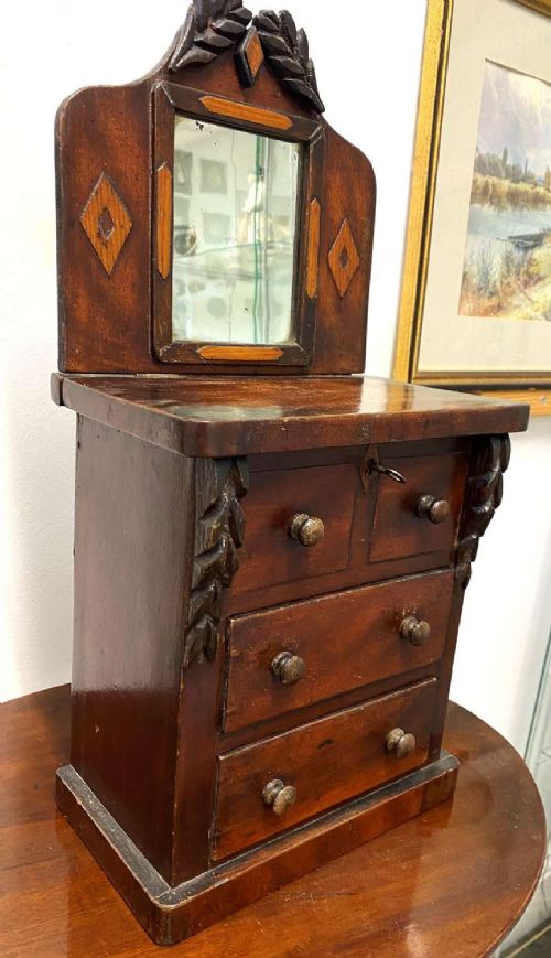A Victorian miniature mahogany chiffonier featuring a small rectangular mirror, carved leaf cresting, inlaid diamond motifs, and small drawers with turned knobs. View V