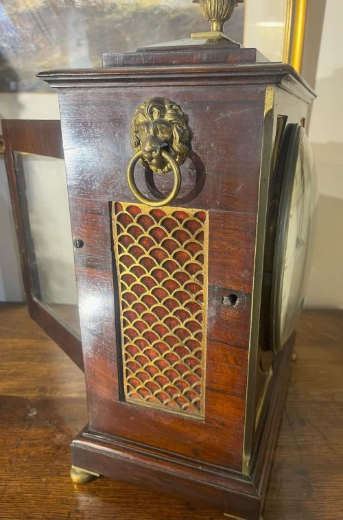 A Regency mahogany bracket clock featuring brass corner inlay, a top pineapple finial, brass ball feet, and a cream painted dial signed P. Ketterer Stamford Hill. View VI