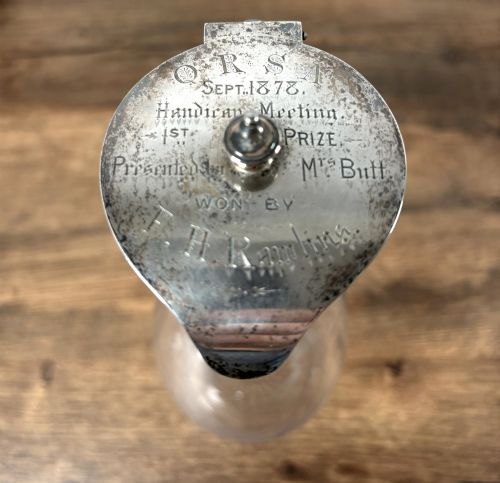 fig 1. A clear glass claret jug featuring a starburst cut base, a loop glass handle, and an open hinged silver lid.