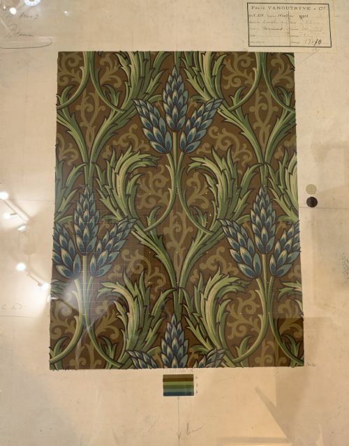 fig 1. A framed antique French textile design on paper featuring stylized green leaves and blue botanical buds against a patterned brown background.