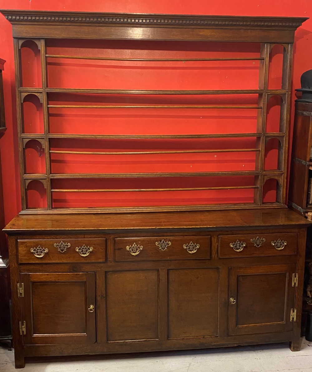 A George III oak dresser featuring an open upper plate rack with slender horizontal rails above three drawers and lower panelled cupboards with brass fittings.