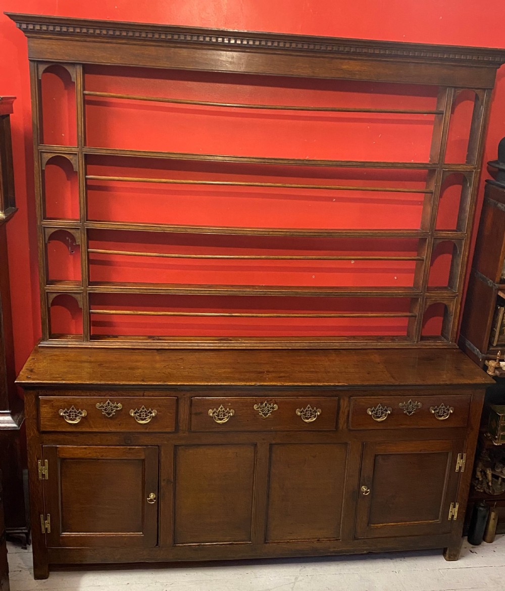 A George III oak dresser featuring an open upper plate rack with slender horizontal rails above three drawers and lower panelled cupboards with brass fittings. View II