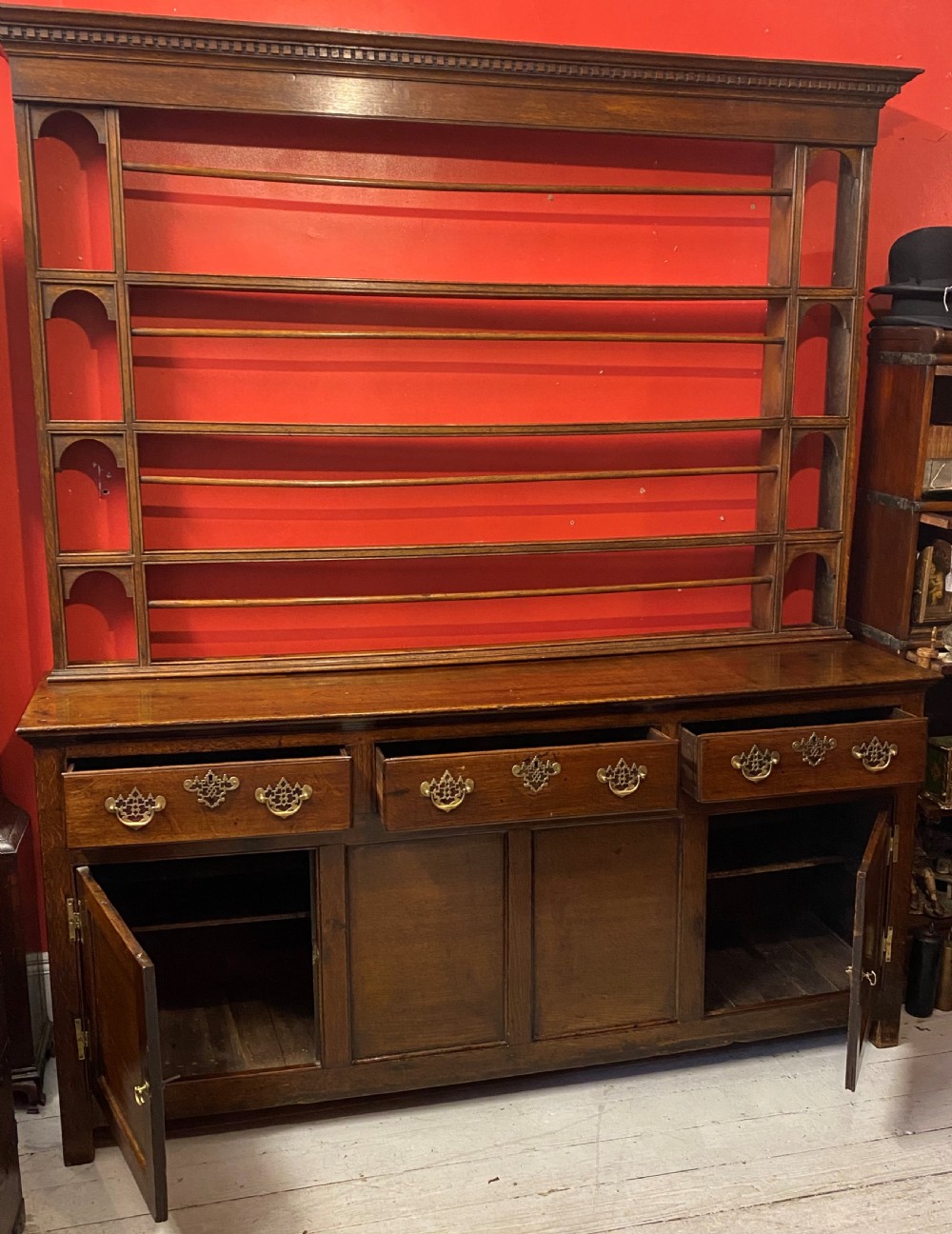 A George III oak dresser featuring an open upper plate rack with slender horizontal rails above three drawers and lower panelled cupboards with brass fittings. View IV