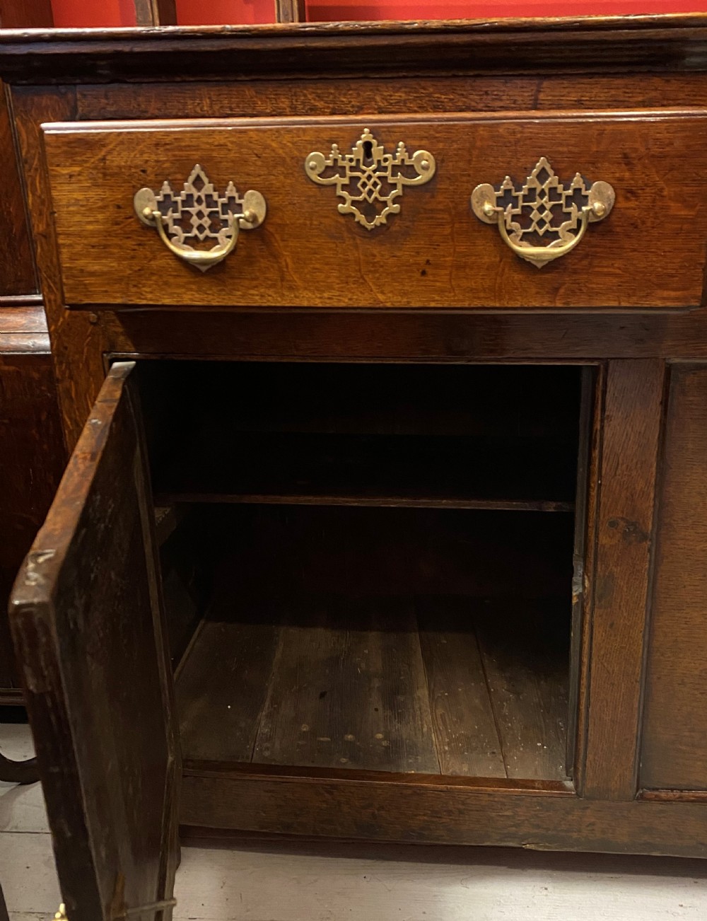 A George III oak dresser featuring an open upper plate rack with slender horizontal rails above three drawers and lower panelled cupboards with brass fittings. View V