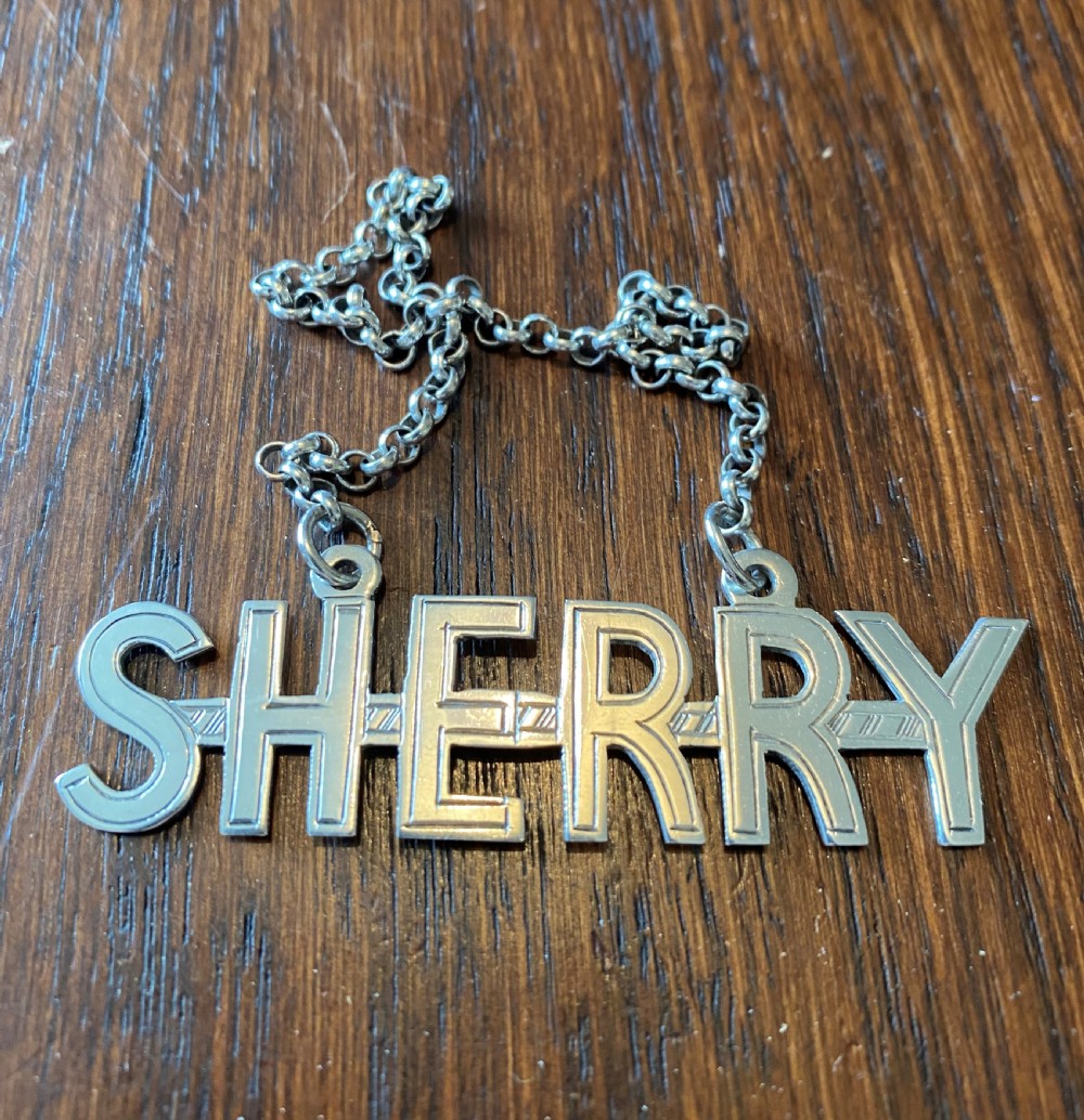 A solid silver decanter label formed from cut-out capital letters spelling SHERRY attached to a fine metal chain.