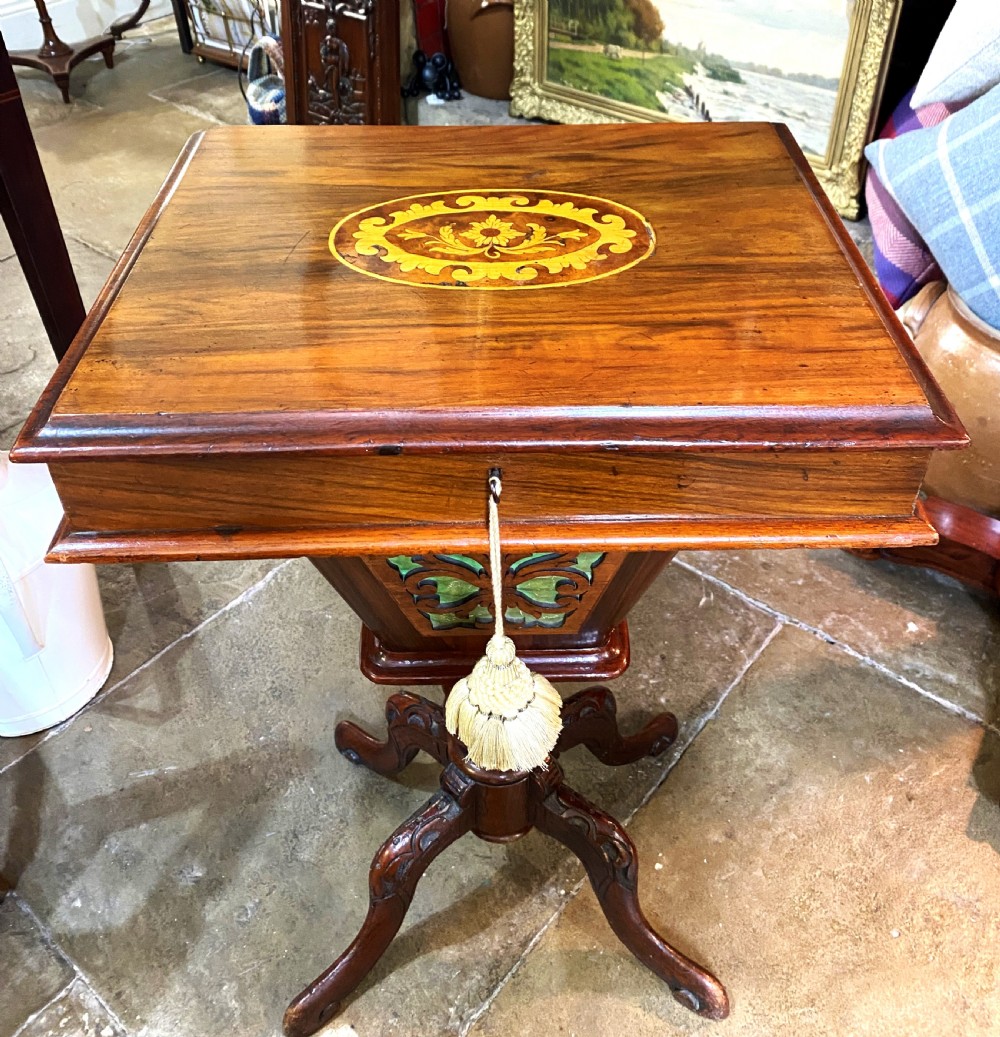 A Victorian walnut sewing table featuring a hinged lid decorated with an oval floral marquetry inlay set upon a carved tripod pedestal base.