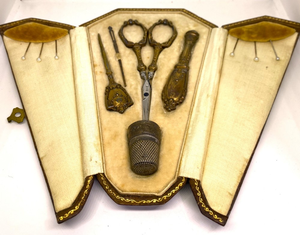 A tri-fold brown leather sewing etui open to display a cream silk interior holding gilt brass scissors, a thimble, and needlework tools.