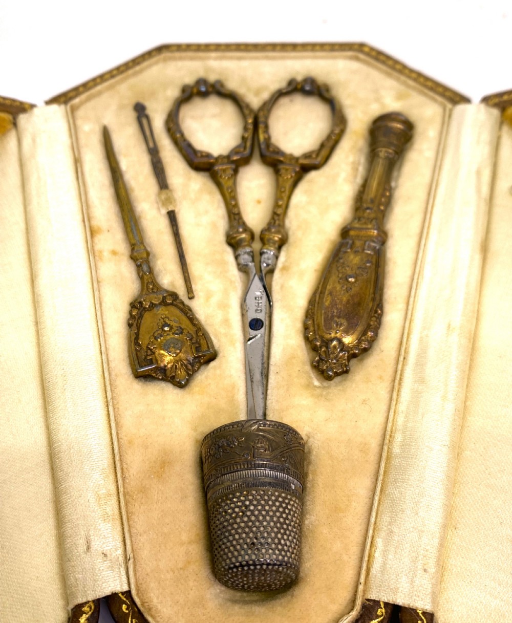 A tri-fold brown leather sewing etui open to display a cream silk interior holding gilt brass scissors, a thimble, and needlework tools. View II