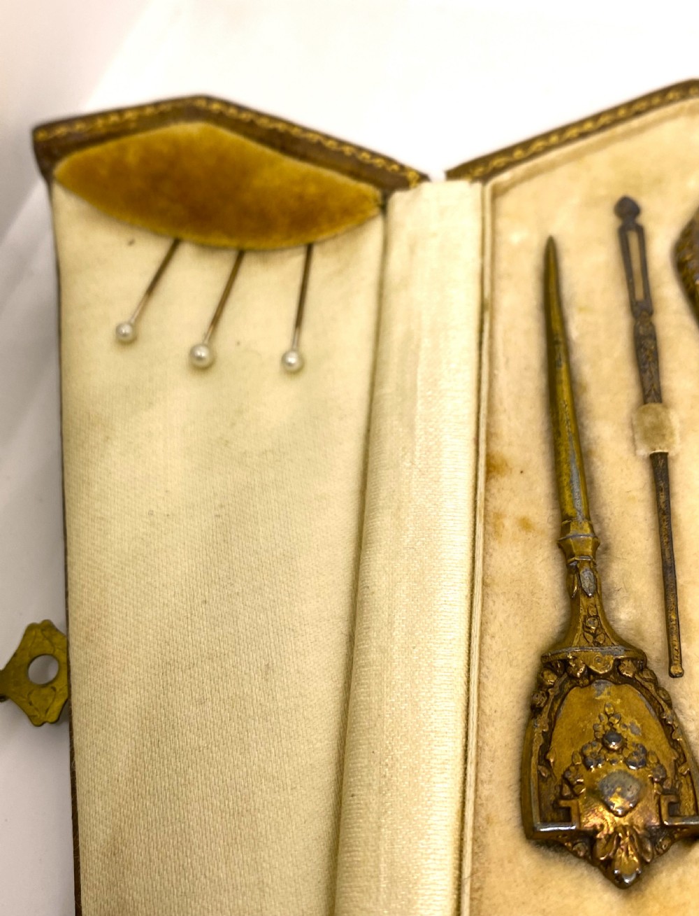 A tri-fold brown leather sewing etui open to display a cream silk interior holding gilt brass scissors, a thimble, and needlework tools. View III