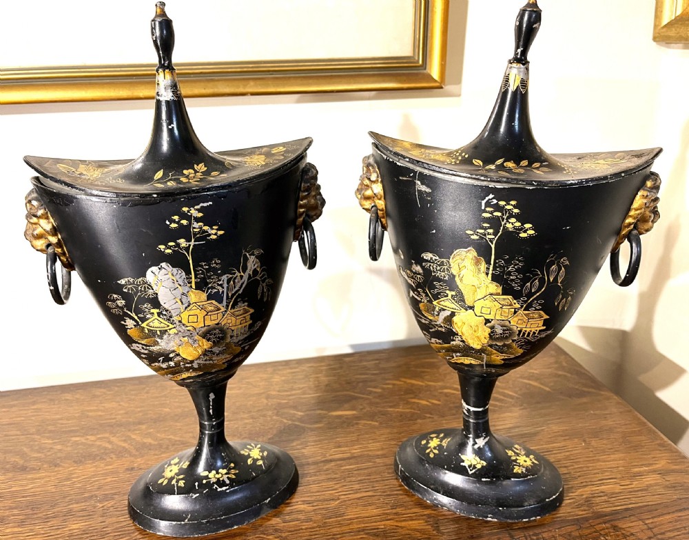 A pair of black painted metal toleware chestnut urns decorated with gold chinoiserie landscape scenes, pointed lids, and lion mask side handles.