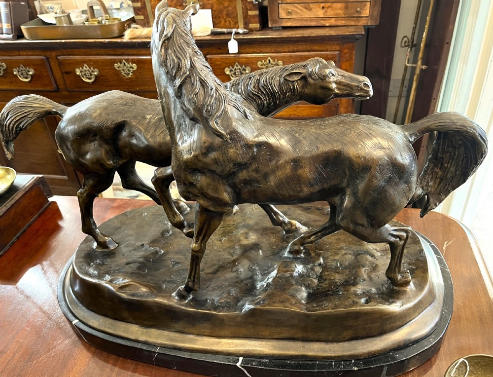 A bronze sculpture depicting two horses standing together on a naturalistic ground base mounted on an oval dark marble plinth.