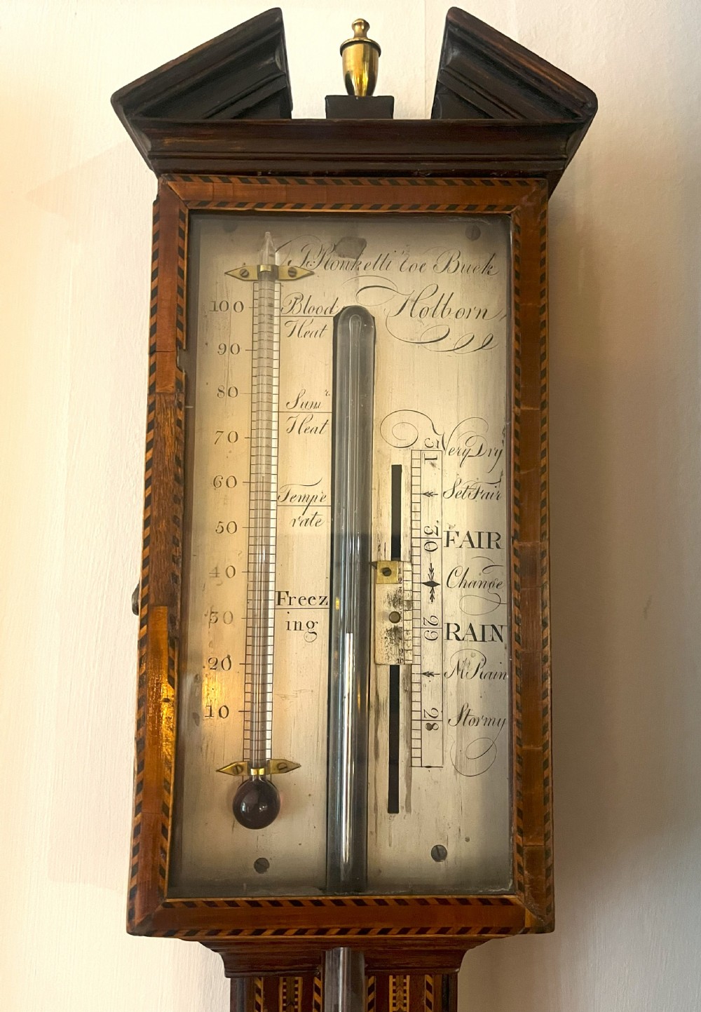 An antique Georgian mahogany stick barometer featuring geometric parquetry inlay, a broken pediment top, and an engraved silvered register plate. View X