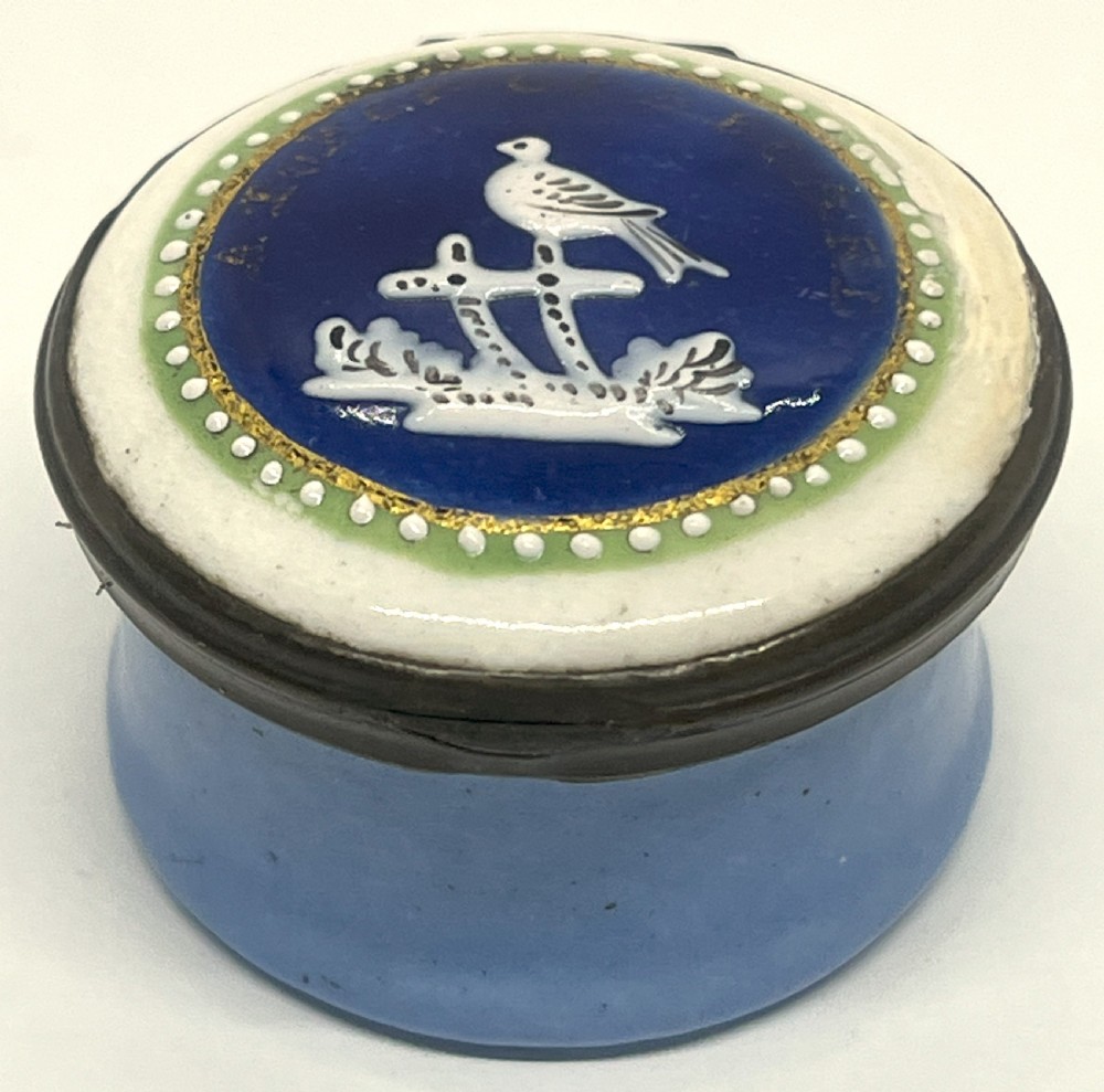 A circular Georgian South Staffordshire enamel patch box featuring a light blue base and a hinged lid adorned with a white bird motif on a navy blue ground.