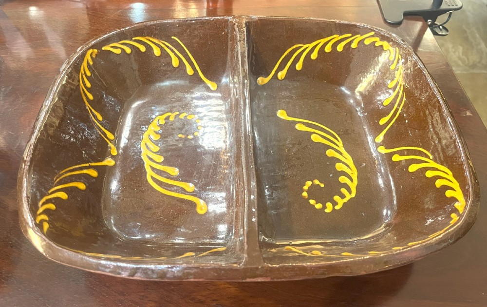 A dark brown glazed earthenware serving dish divided into two sections, featuring yellow trailed slip feather motifs along the interior.