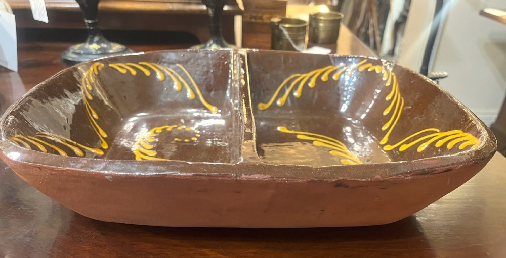 A dark brown glazed earthenware serving dish divided into two sections, featuring yellow trailed slip feather motifs along the interior. View II