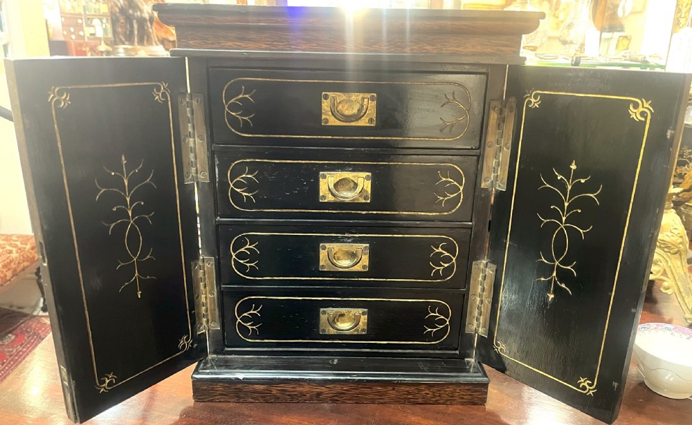A nineteenth-century ebonised palmwood table cabinet with opened double doors revealing four small drawers with flush brass handles and gold foliate decoration.