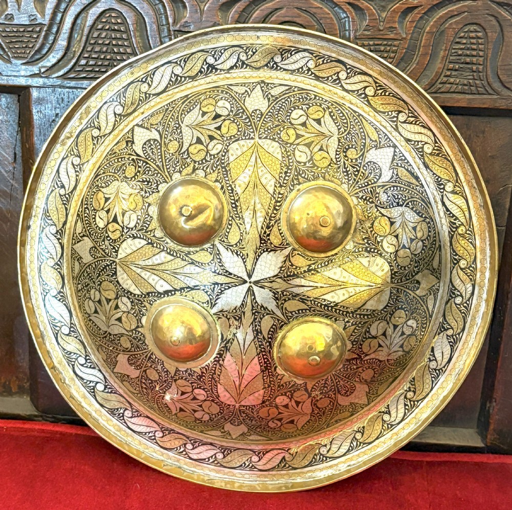 A convex circular metal dhal shield decorated with detailed foliate engraving in gold and silver tones and featuring four prominent raised brass bosses.