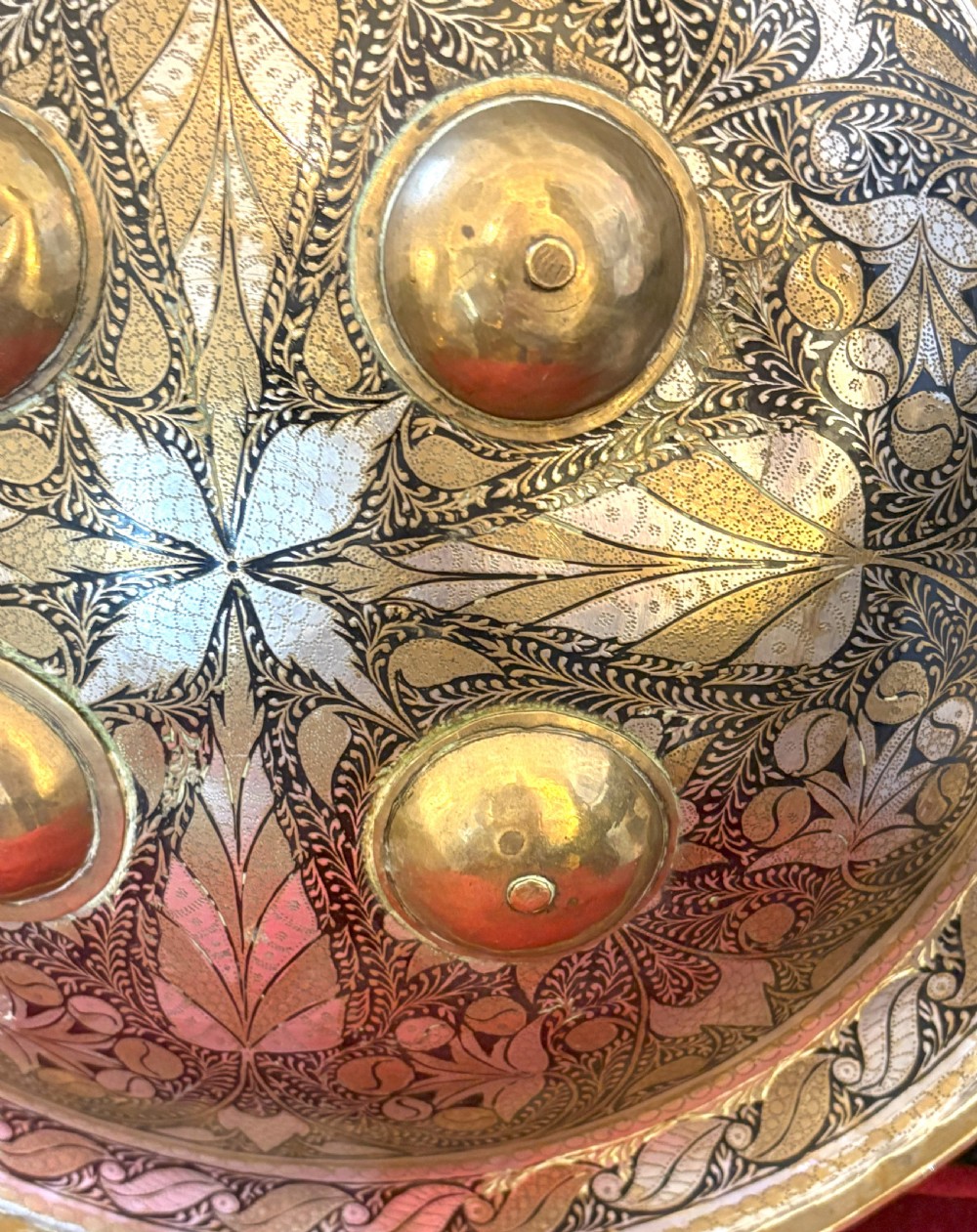 A convex circular metal dhal shield decorated with detailed foliate engraving in gold and silver tones and featuring four prominent raised brass bosses. View II