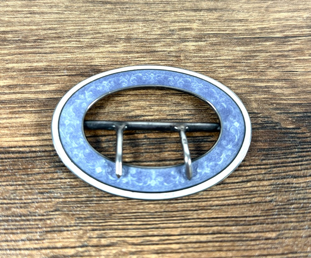 An oval silver belt buckle featuring a light blue guilloche enamel border with foliate scroll engraving and a double central prong.