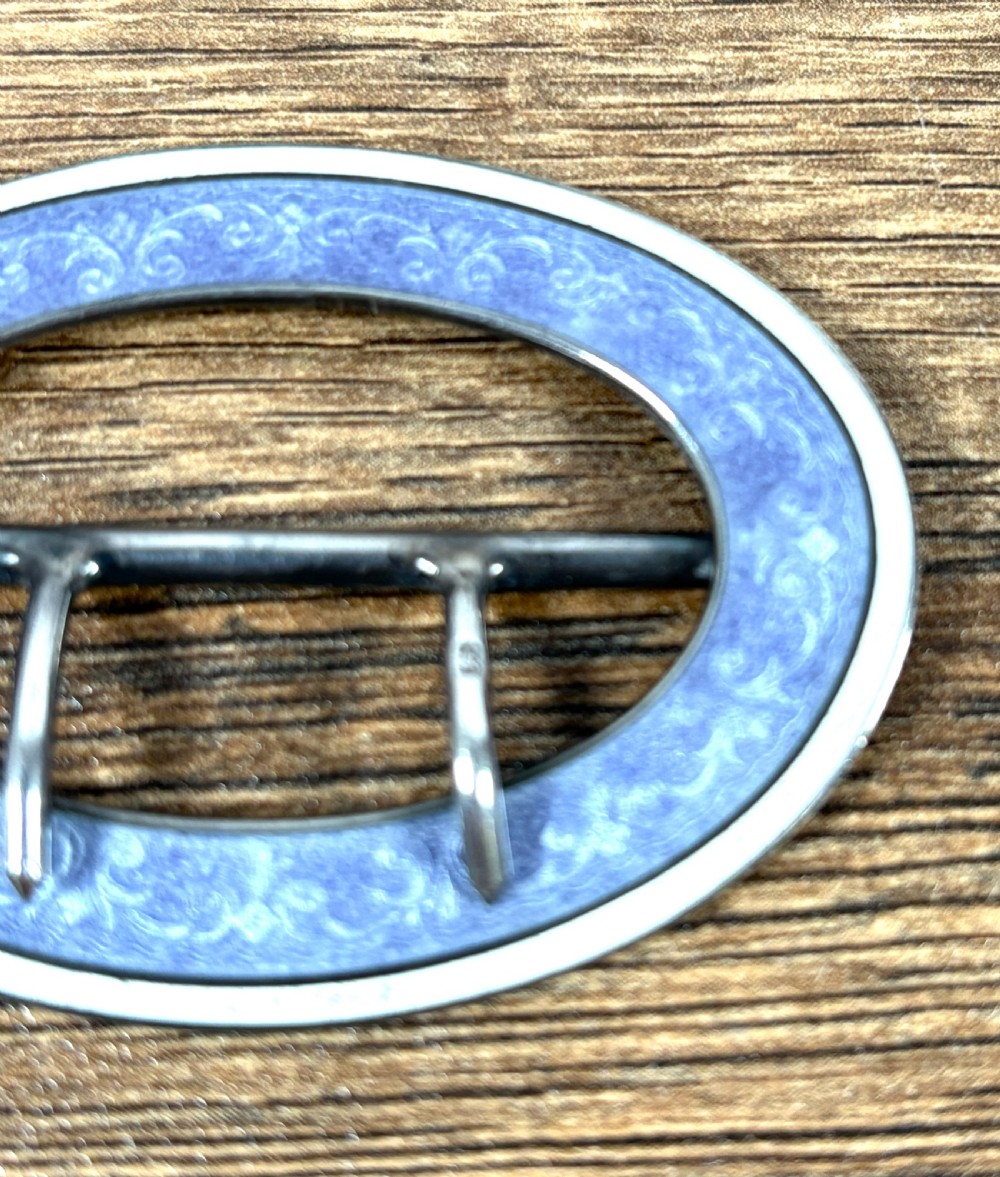 An oval silver belt buckle featuring a light blue guilloche enamel border with foliate scroll engraving and a double central prong. View II