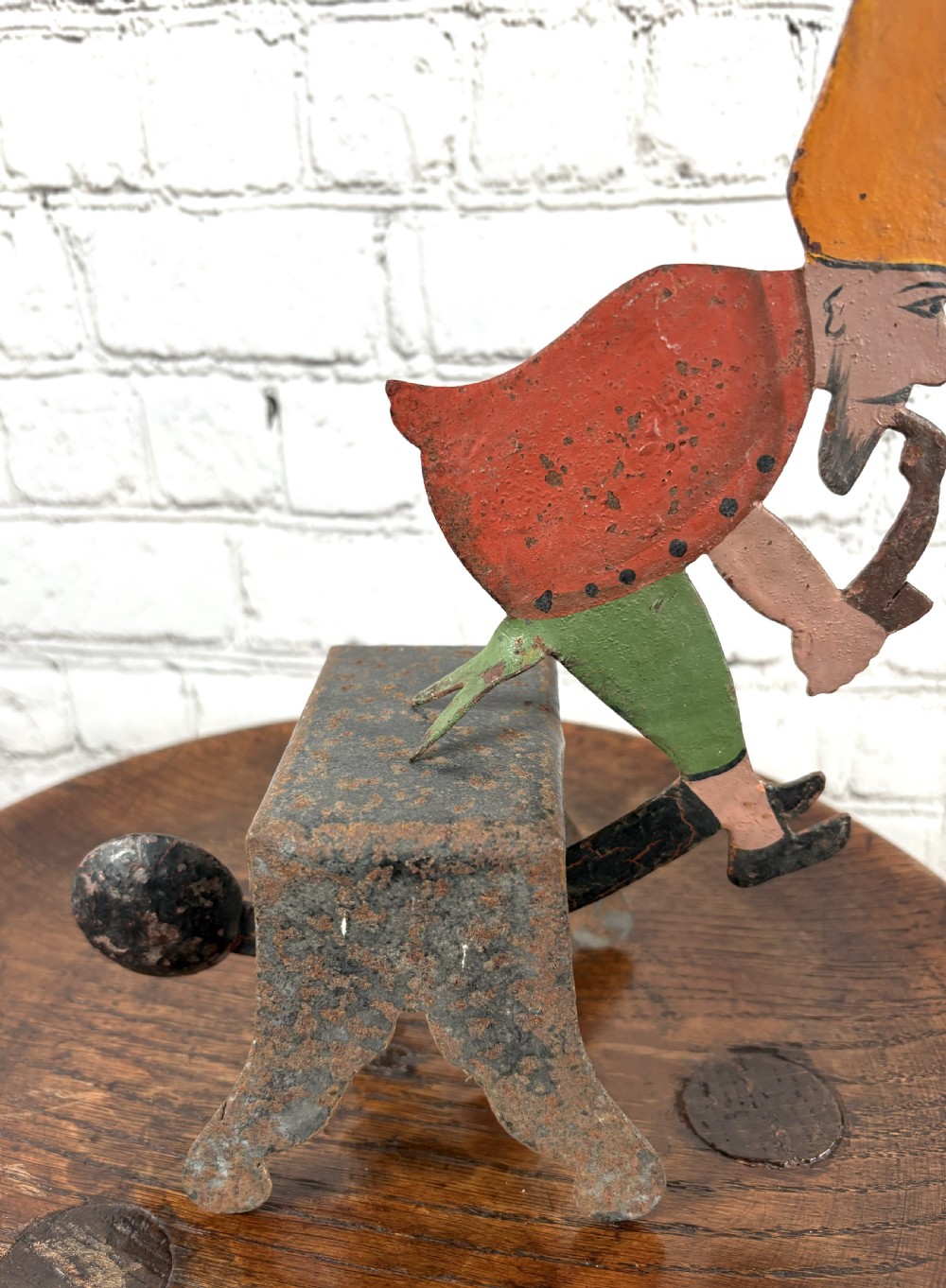 A painted metal balance toy depicting a figure in an orange hat smoking a pipe, mounted on an iron base with a counterweight. View II