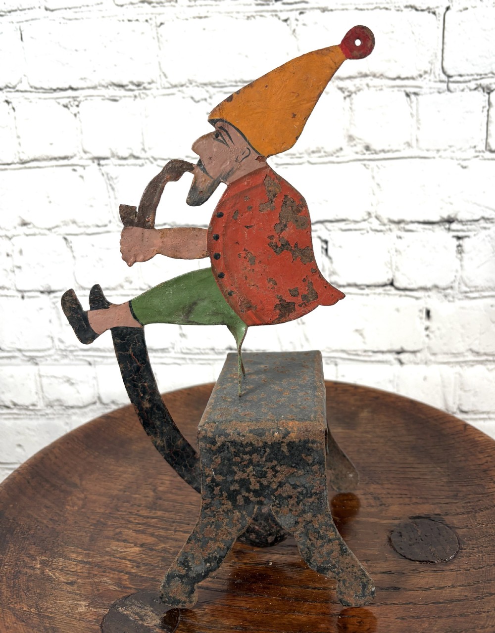 A painted metal balance toy depicting a figure in an orange hat smoking a pipe, mounted on an iron base with a counterweight. View III