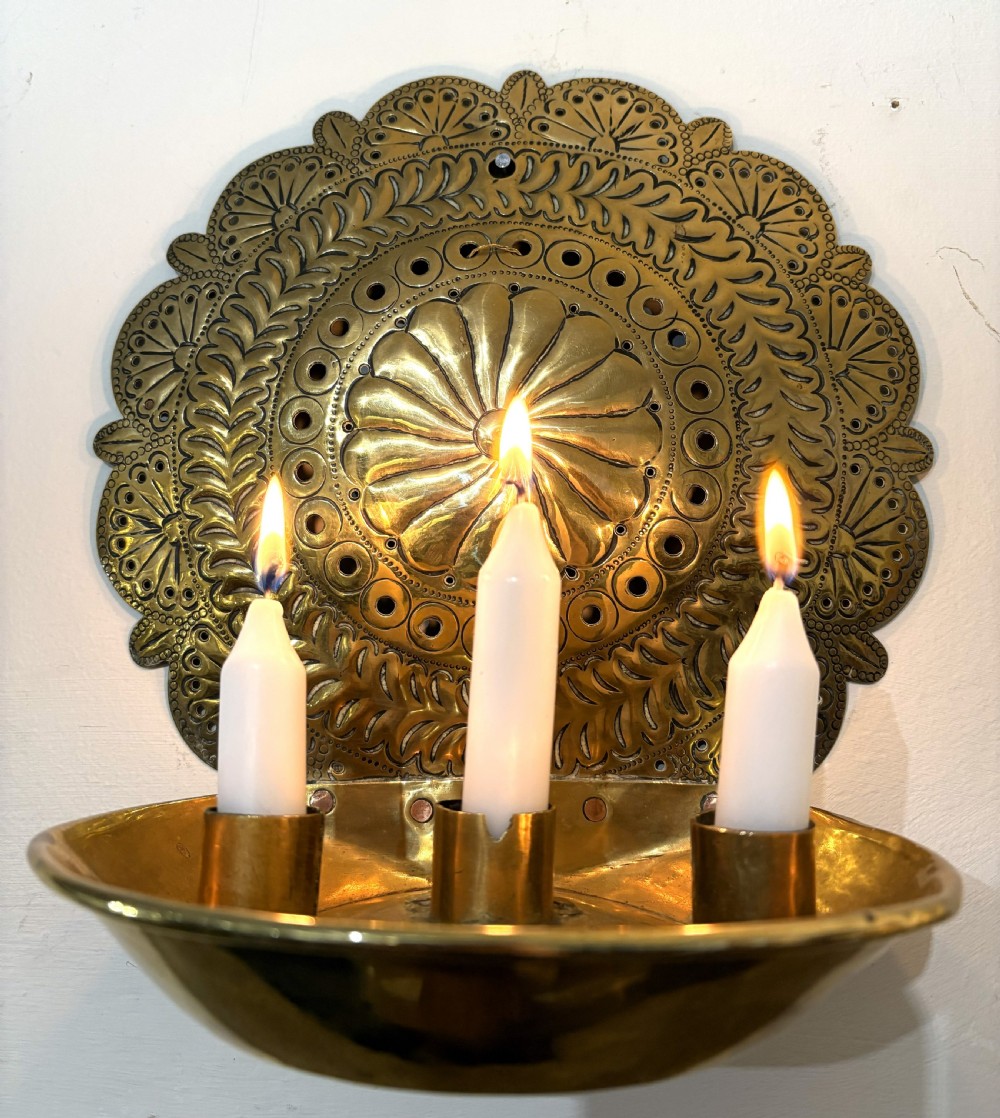 An 18th-century brass wall sconce featuring a round chased backplate with floral motifs and three candle nozzles holding lit white candles.