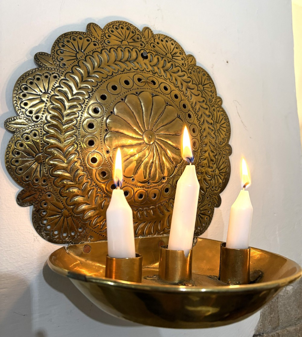 An 18th-century brass wall sconce featuring a round chased backplate with floral motifs and three candle nozzles holding lit white candles. View II