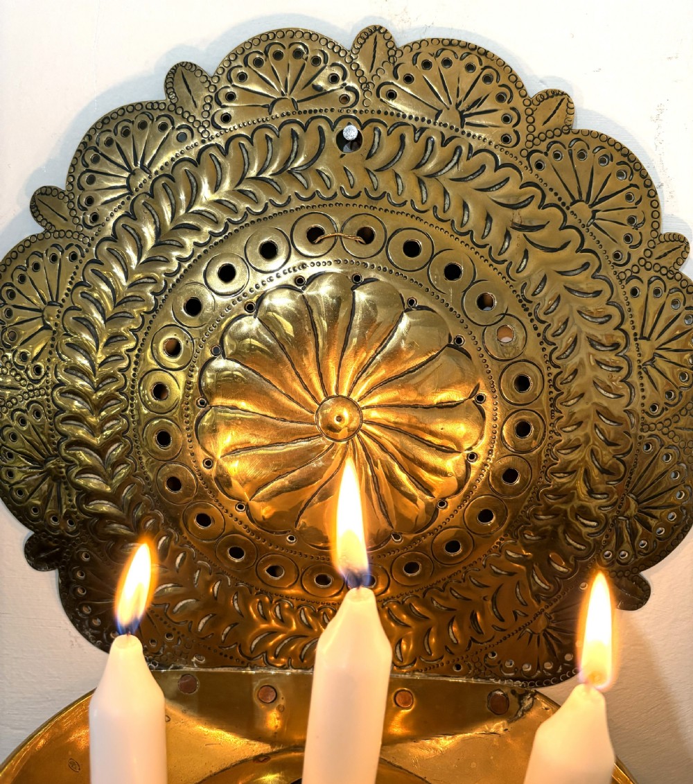 An 18th-century brass wall sconce featuring a round chased backplate with floral motifs and three candle nozzles holding lit white candles. View III