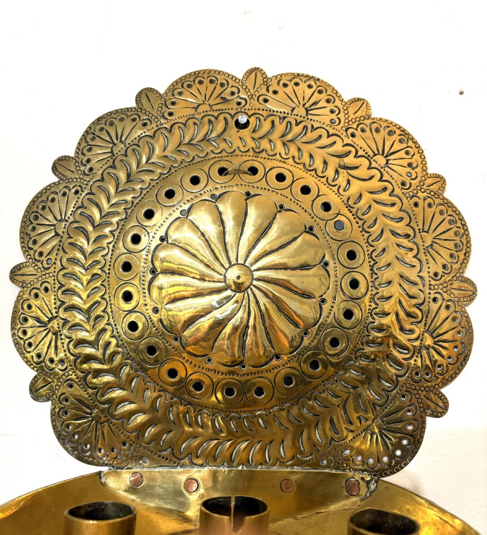 An 18th-century brass wall sconce featuring a round chased backplate with floral motifs and three candle nozzles holding lit white candles. View IV