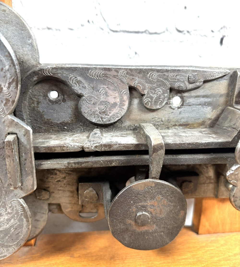 A 17th-century French wrought iron twin spring bolt door lock featuring detailed foliate scroll engravings and lobed metal plates, mounted on a light timber display stand. View III